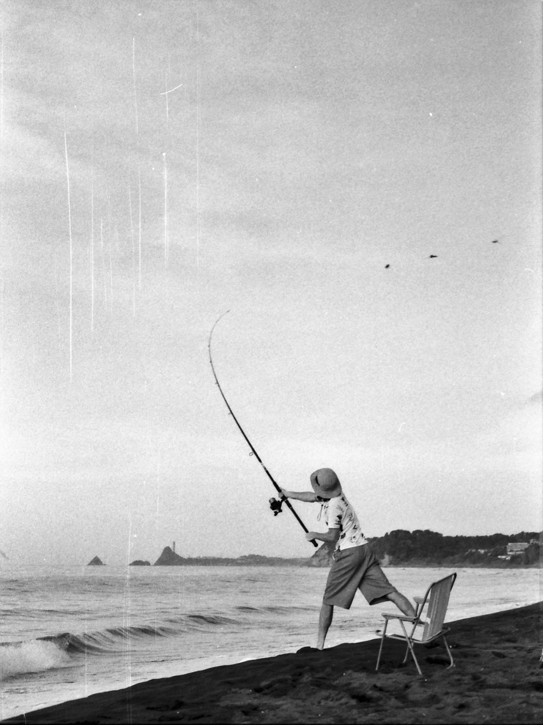 A person wearing a hat and shorts casts a fishing rod into the ocean from a dark sand beach.