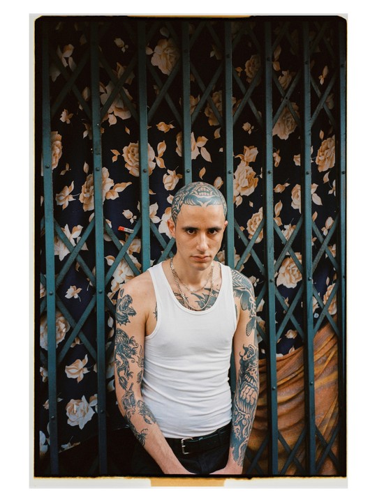 A man with tattoos on his head and arms stands in front of a floral curtain and a metal gate.