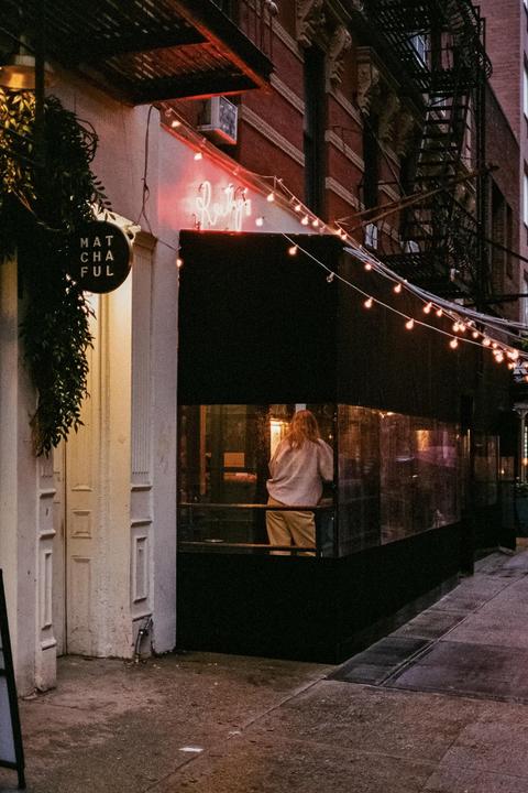 A woman stands inside a restaurant with string lights and a neon sign on its exterior.