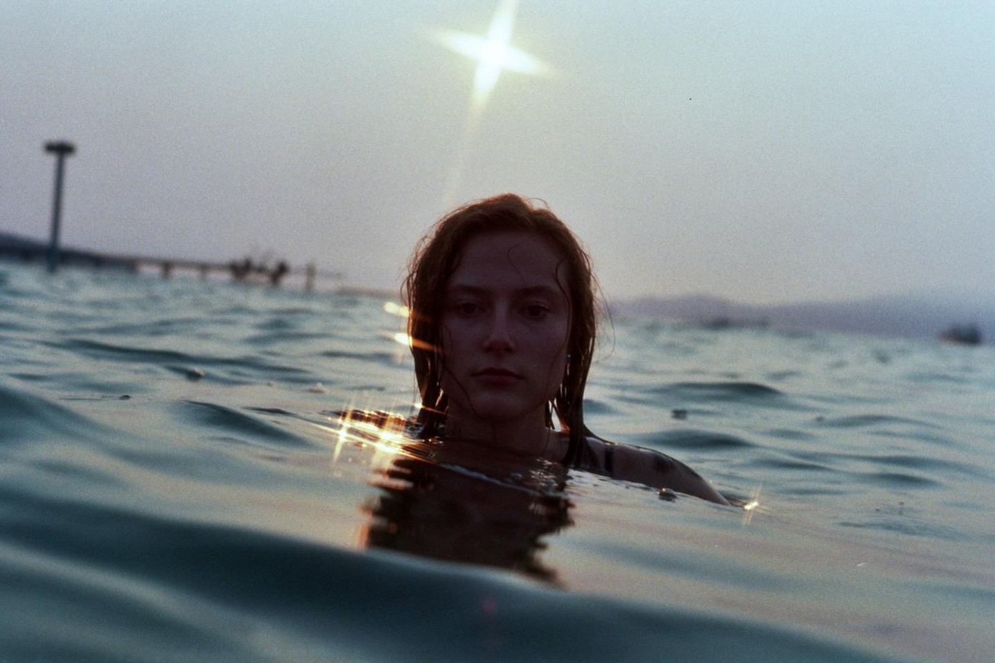 A woman with wet hair is in the water with a bright sun flare above her head.