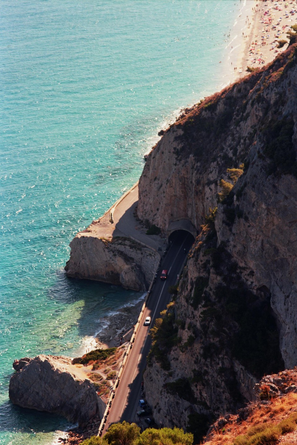 A winding road with a tunnel runs along a steep, rocky coastline next to a turquoise sea, with a beach visible in the distance.
