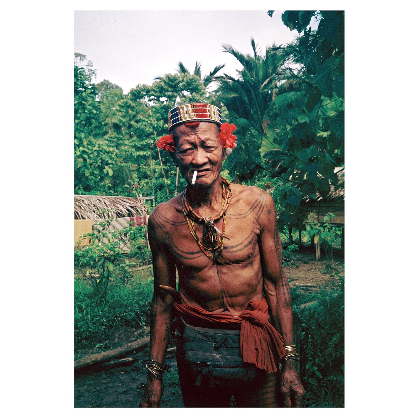 A man with tattoos and a cigarette in his mouth stands in a lush green environment.