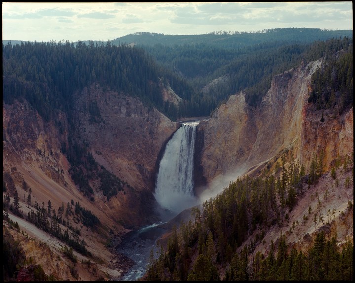 A large waterfall cascades into a river at the bottom of a canyon surrounded by pine trees.
