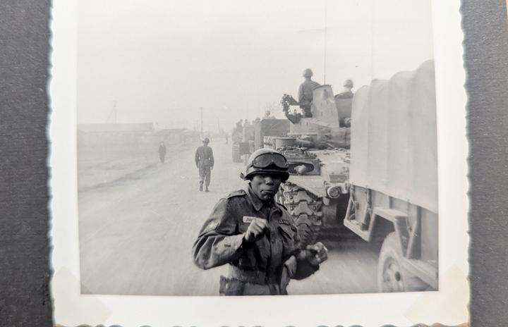 A man in military uniform and goggles stands in the foreground with military vehicles and other soldiers behind him on a hazy day.