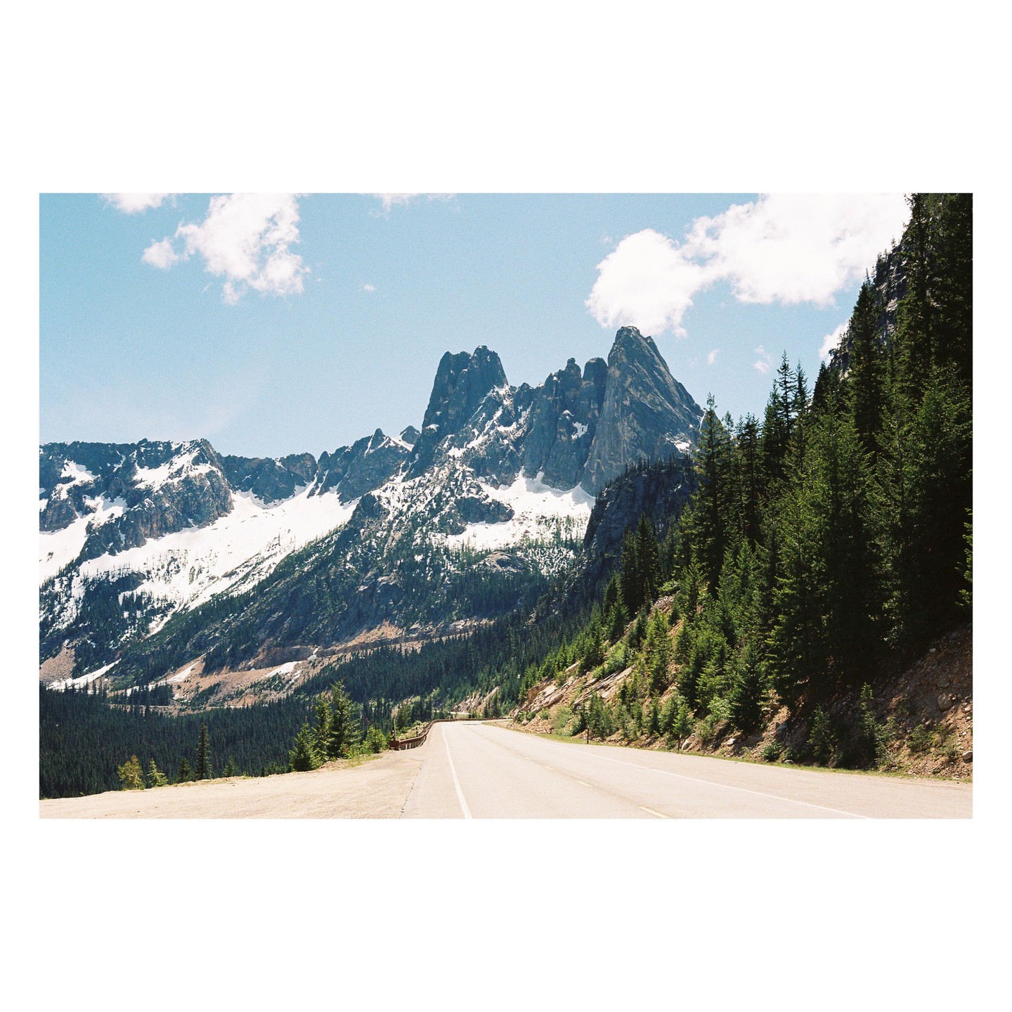 A road winds through a mountainous landscape with snow-capped peaks and evergreen trees under a blue sky with clouds.