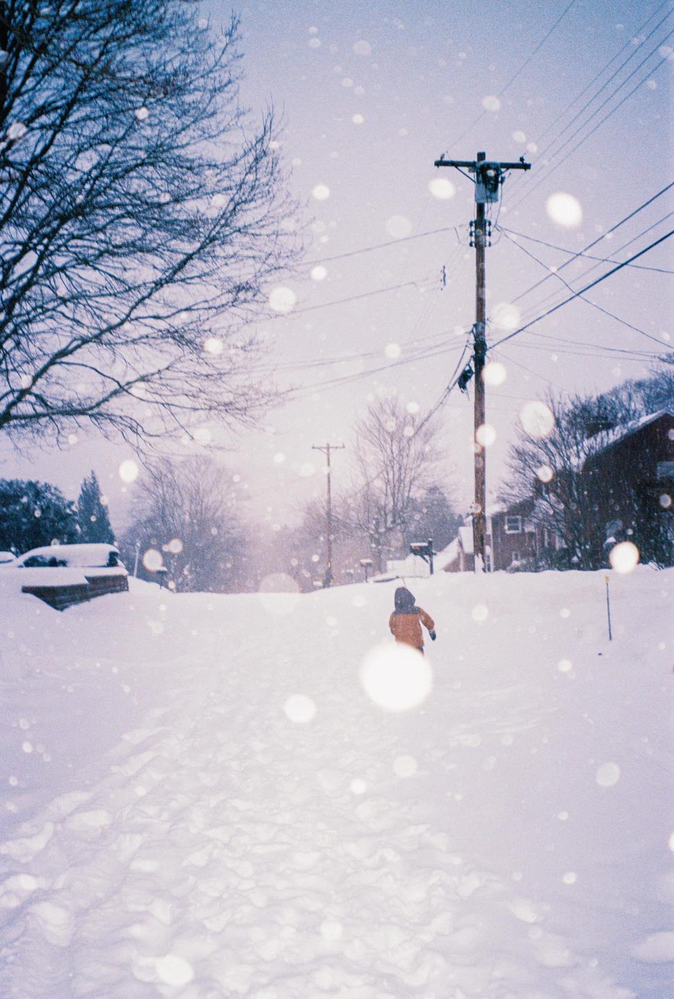 A child in an orange jacket walks away from the viewer through a snow-covered street during a blizzard.