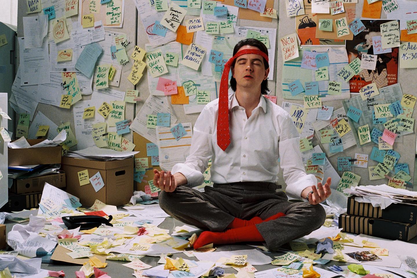 A man sits cross-legged on the floor in a room covered in notes and papers, meditating with his eyes closed.