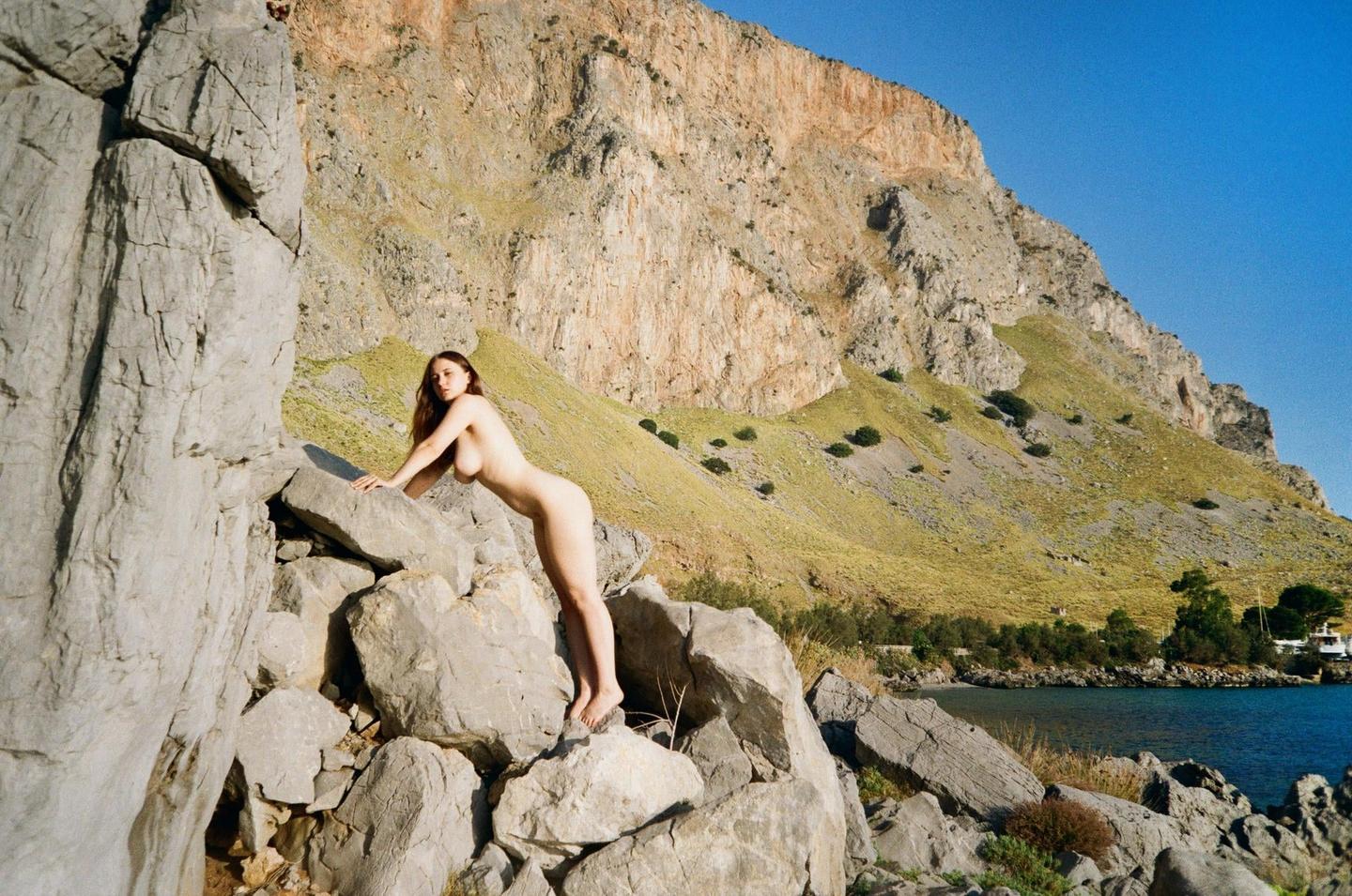 A nude woman poses on rocks by the sea with a large mountain in the background.