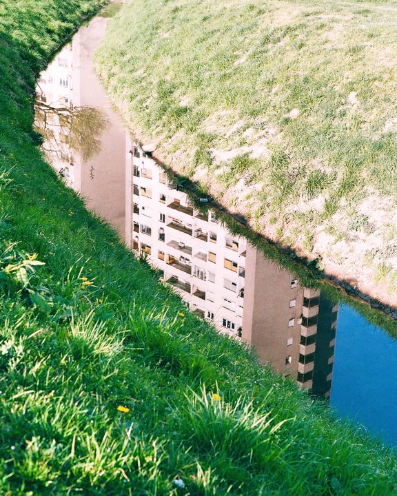 A tall building is reflected in a narrow canal surrounded by grassy banks.