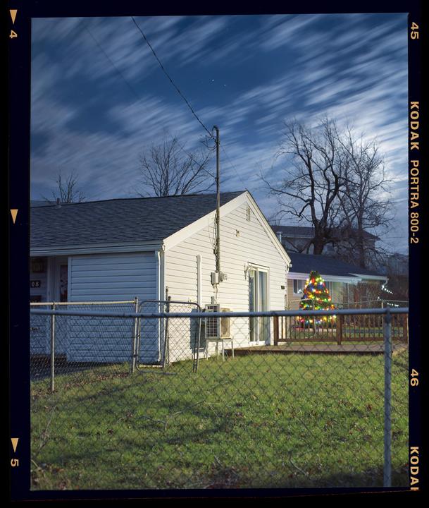 A white house with a chain link fence in the foreground is visible under a cloudy night sky, with a decorated Christmas tree in the background.