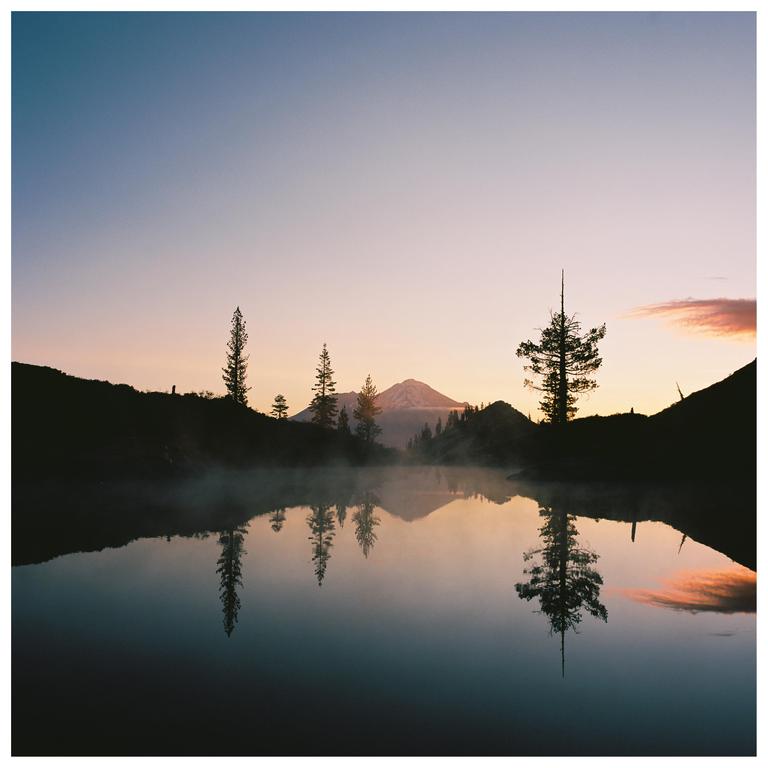 A mountain with a snow-capped peak is reflected in a misty lake at dusk, with silhouetted trees and hills in the foreground.