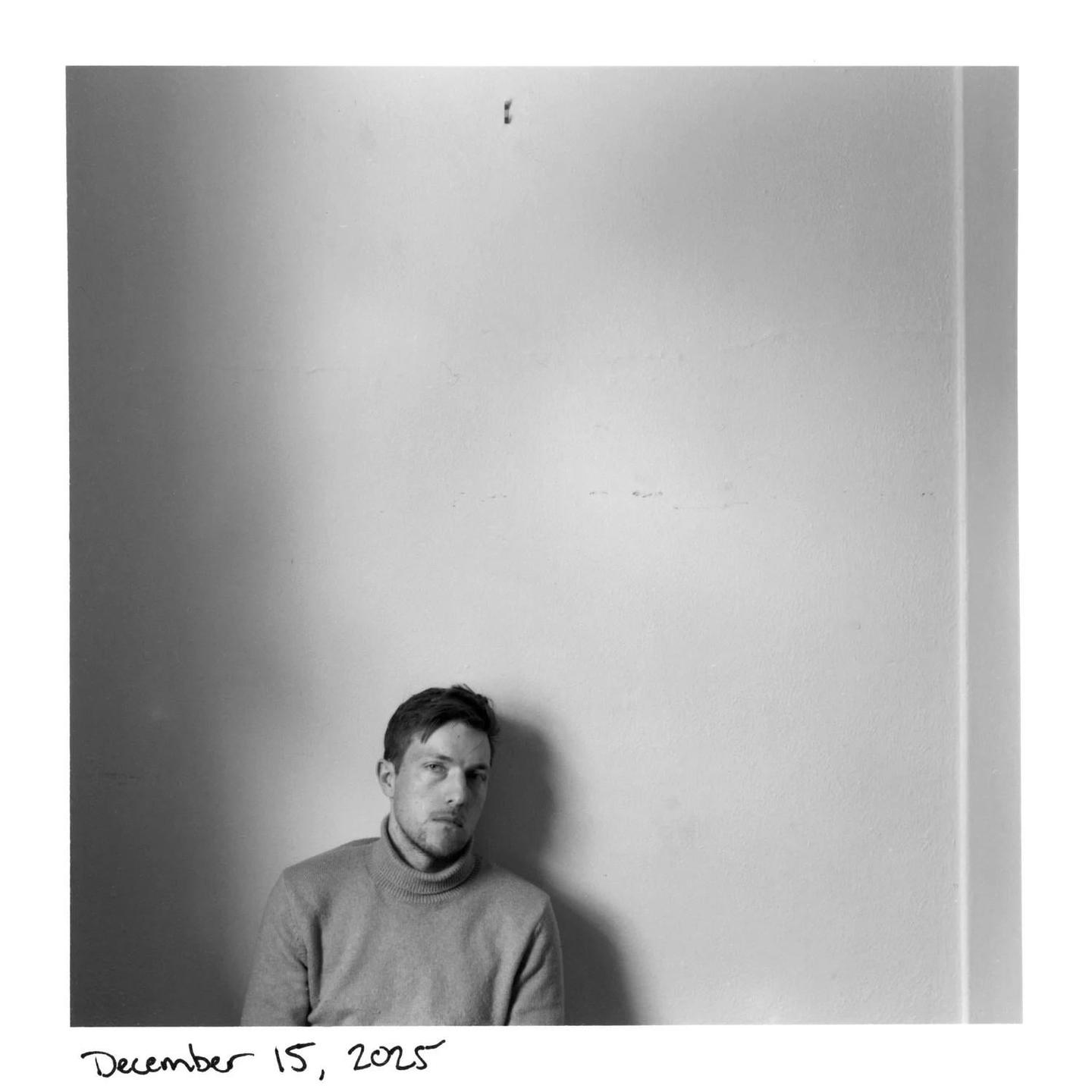 A man in a turtleneck sweater sits against a plain wall.