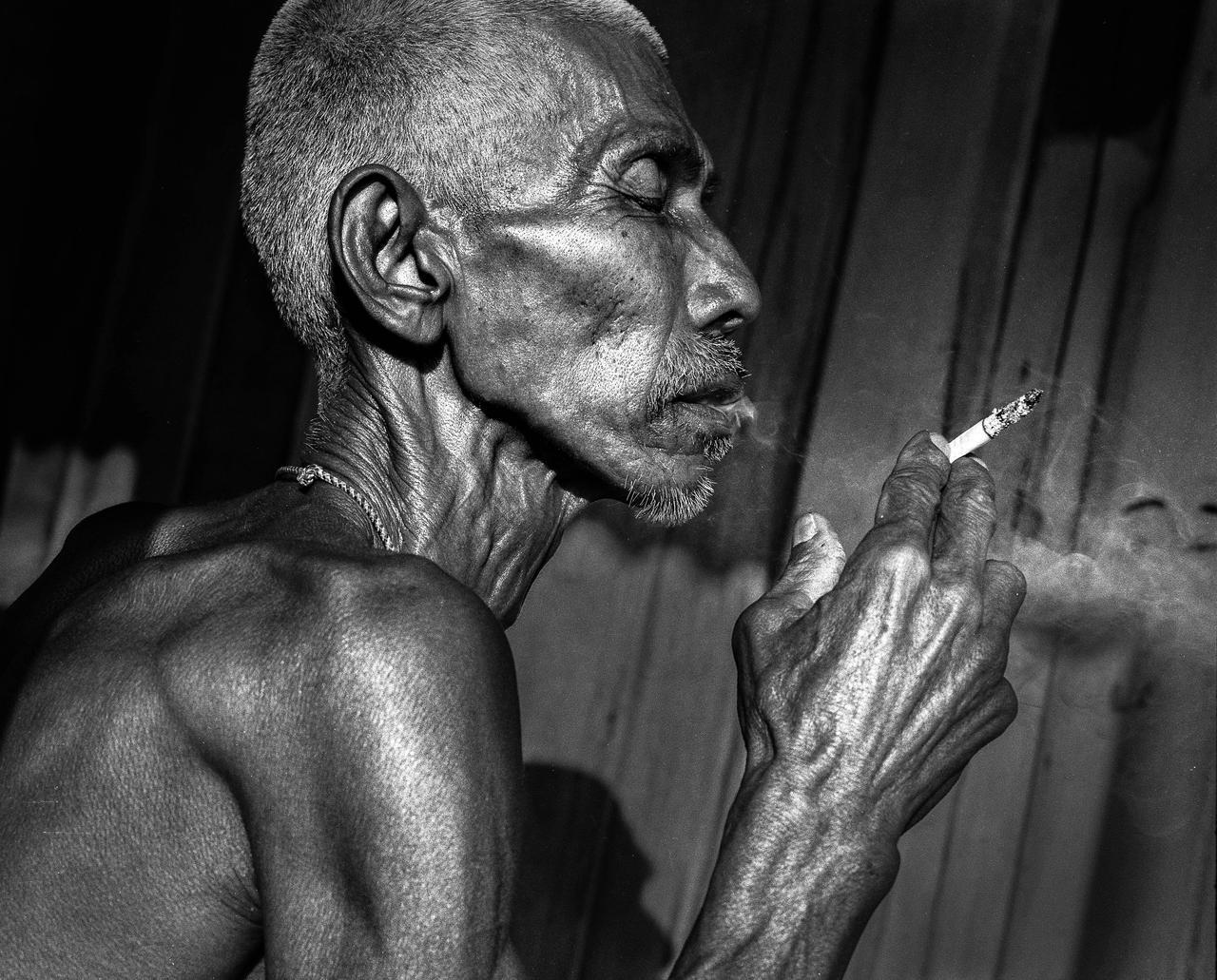 A man with closed eyes holds a lit cigarette, with smoke rising from it.