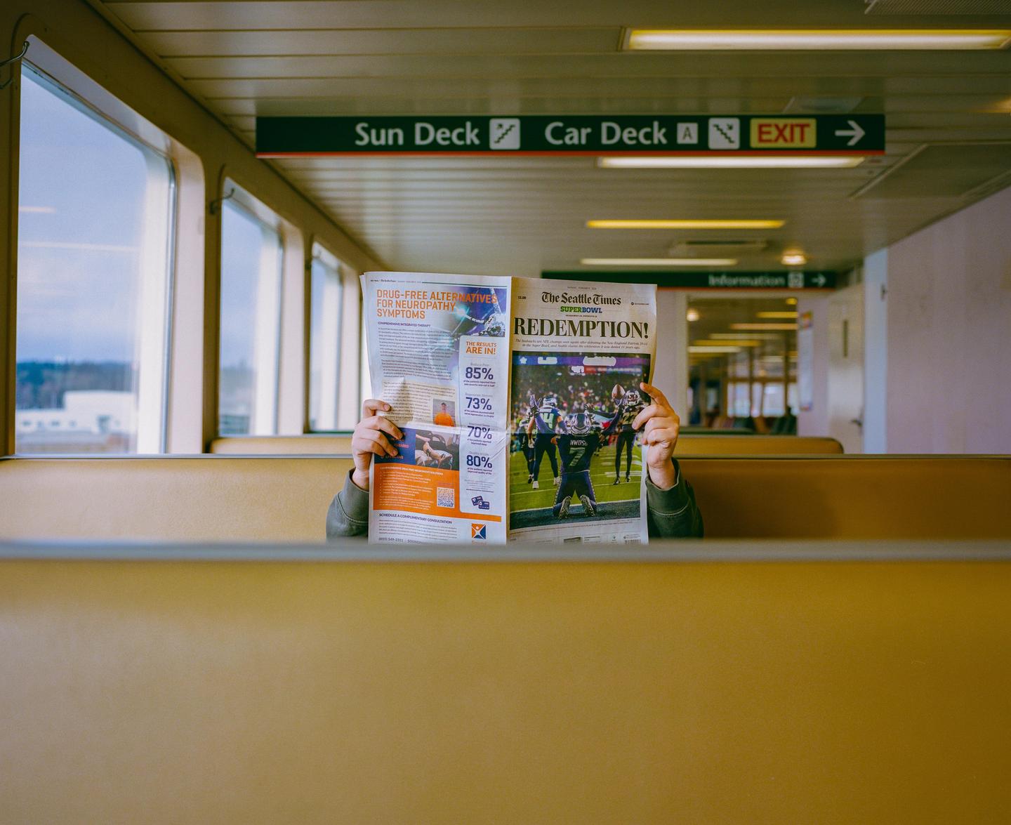 A person holds up a newspaper, obscuring their face, while sitting on a ferry.