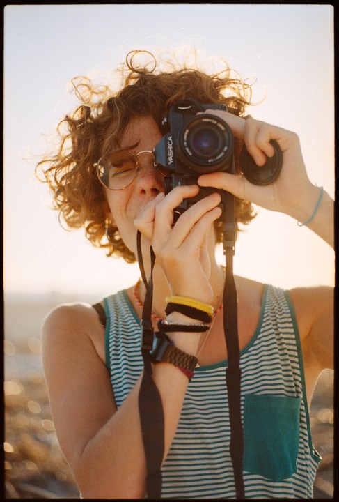 A woman with curly hair and glasses holds a camera up to her face, taking a self-portrait.