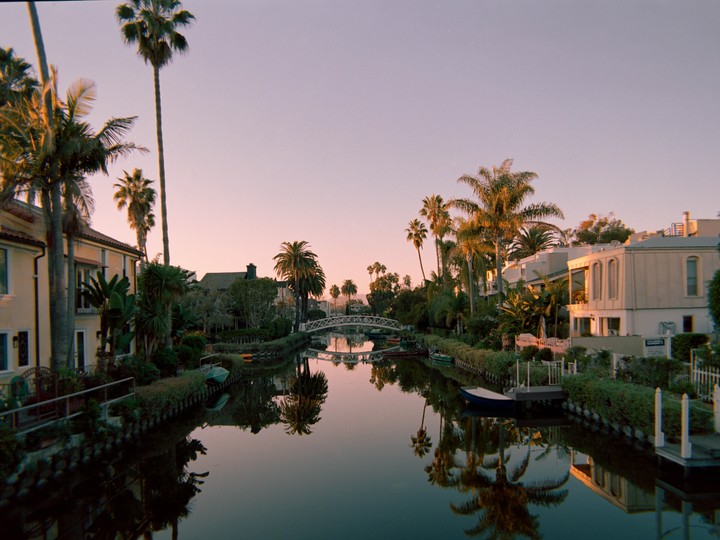 A canal lined with houses and palm trees reflects in the water under a pink sky at sunset.