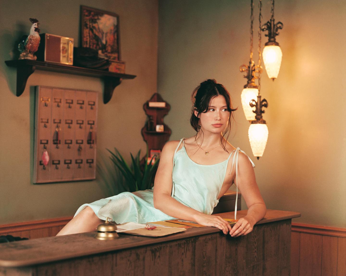 A woman in a light blue dress is leaning on a wooden counter with a bell, next to a key rack and hanging lamps.