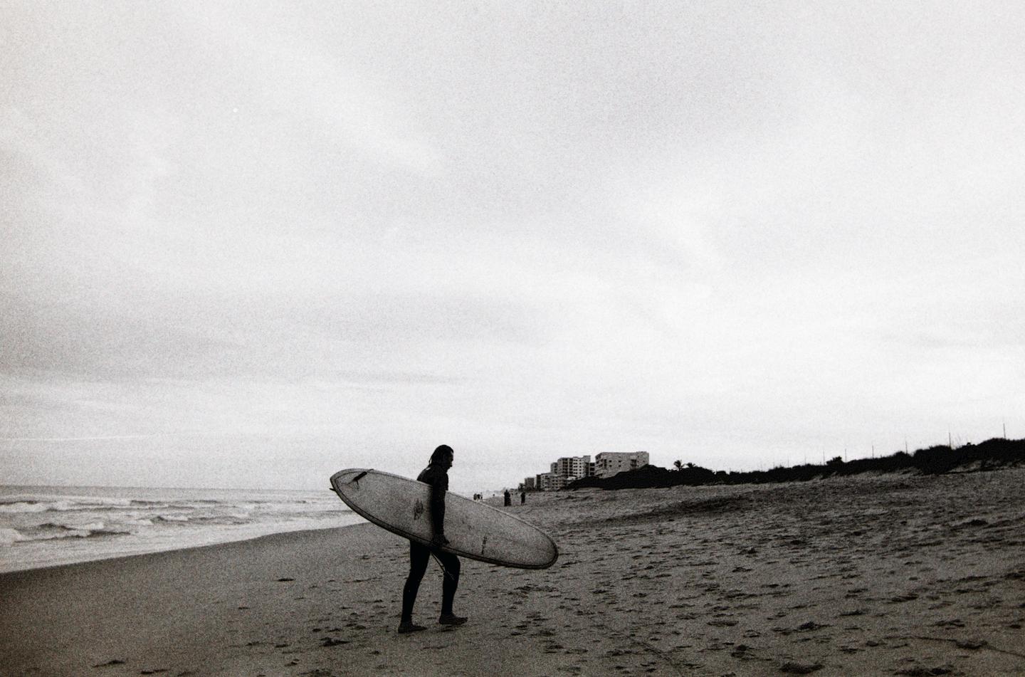 A surfer carrying a surfboard walks along a sandy beach with waves crashing on the left and buildings in the distance on the right.