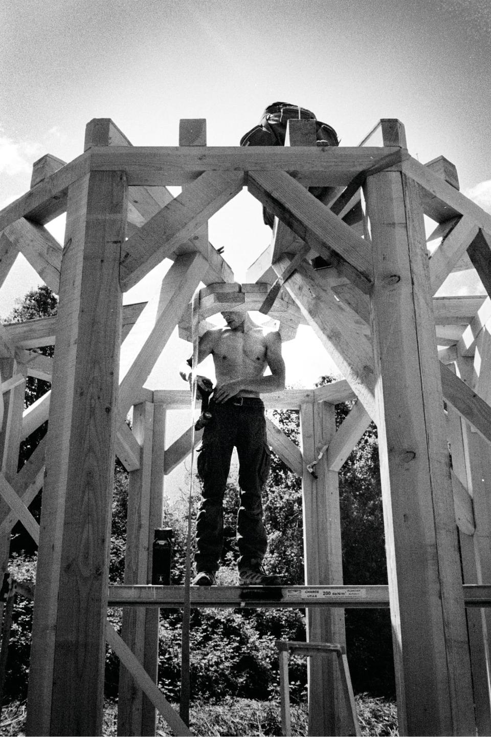 A shirtless man stands on a wooden structure, holding a tool, with trees visible in the background.
