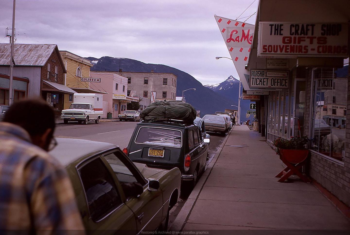 A man stands on a street with parked cars and buildings, with mountains in the background.