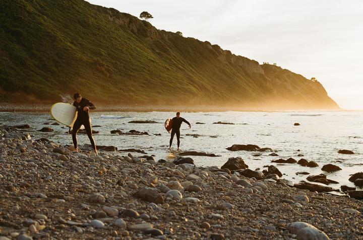 Two men in wetsuits with surfboards stand on a rocky beach at sunset with a large green hill in the background.