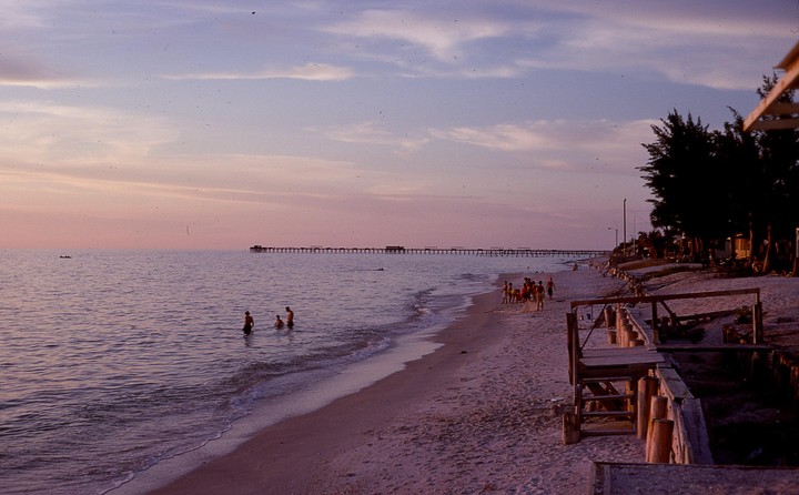A long pier stretches into the ocean at sunset, with people swimming and playing on the beach.