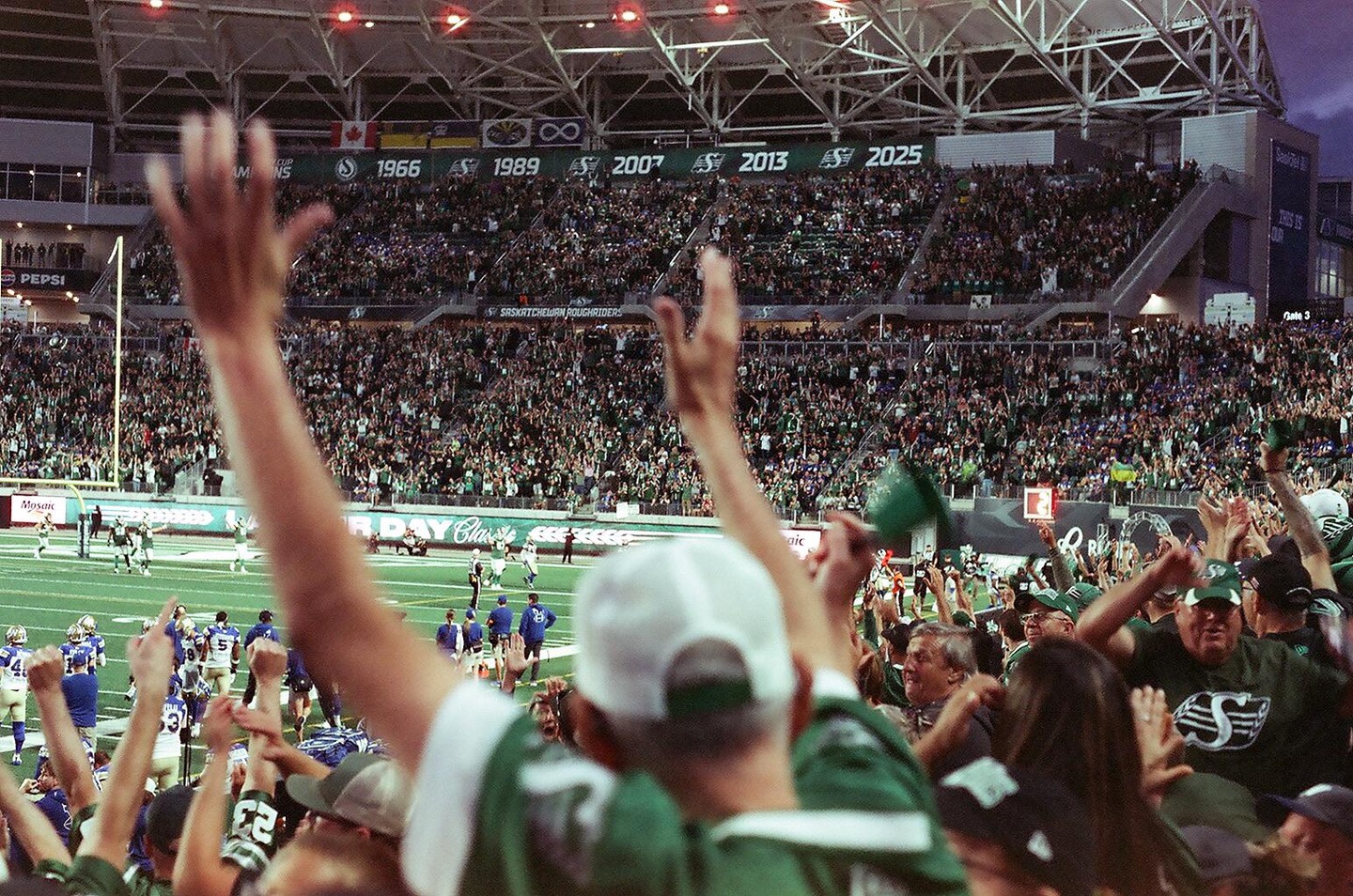 A crowd of people with their arms raised watches a football game in a stadium.