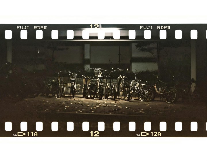 A row of bicycles is parked on a paved surface at night, illuminated by an overhead light.