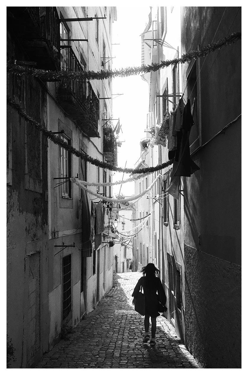 A woman walks down a narrow, cobbled alleyway lined with buildings and strung with garlands and laundry.