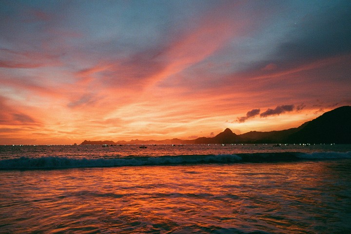 The ocean reflects the vibrant orange and red hues of a sunset sky, with distant mountains and small boats visible on the horizon.
