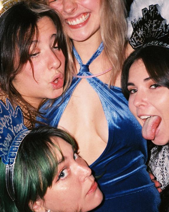 Four women are posing closely together, with one woman in a blue dress at the center.