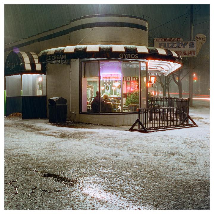 A restaurant with a striped awning and neon signs is covered in snow at night.
