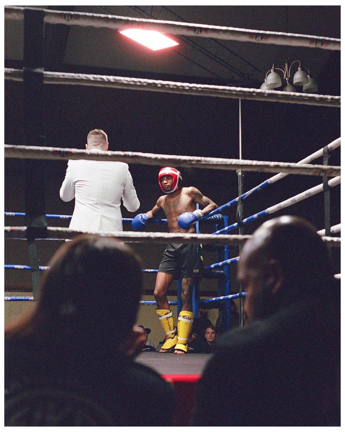A boxer in a red headguard and blue gloves stands in a boxing ring with a man in a white suit.