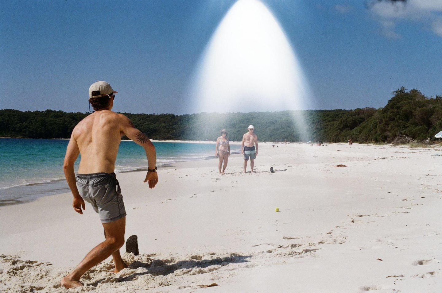 A man in a baseball cap and swim trunks stands on a sandy beach with two people in the distance and a bright light leak across the sky.
