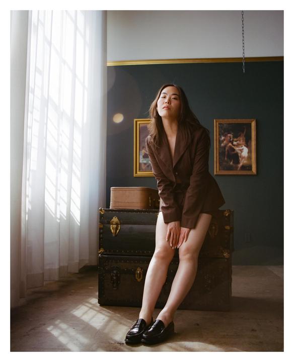 A woman in a brown blazer and loafers sits on stacked trunks in a room with paintings on the wall and light streaming through a window.