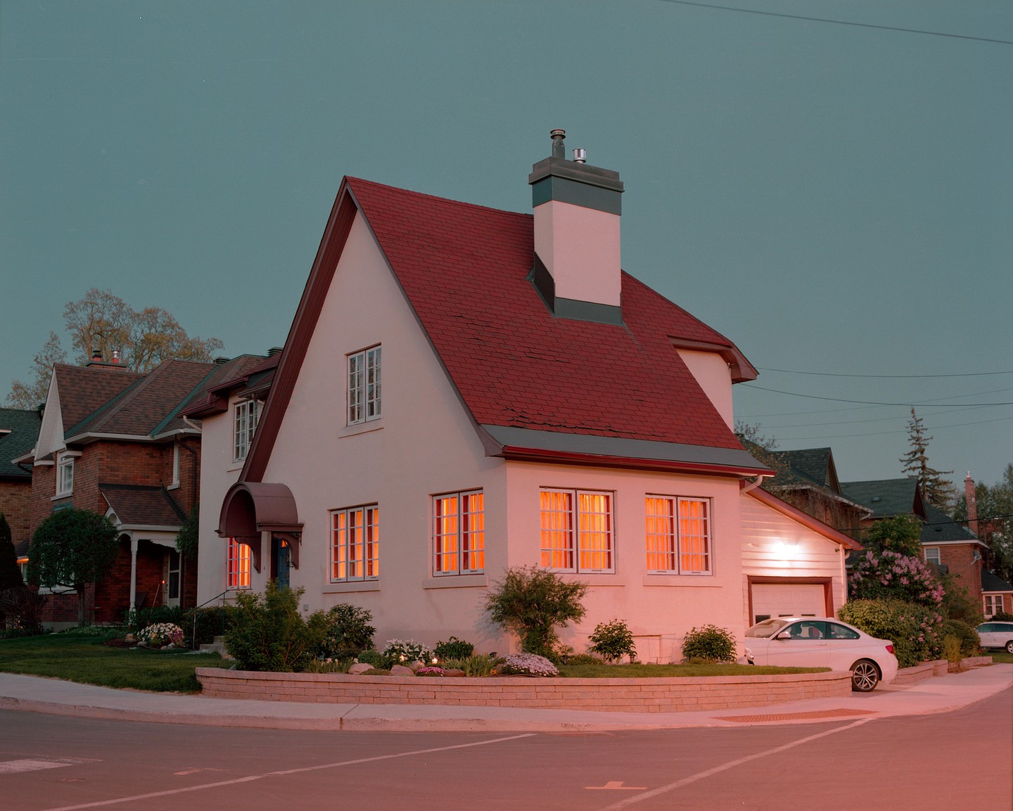 A white house with a red roof and glowing windows sits on a street corner at dusk.