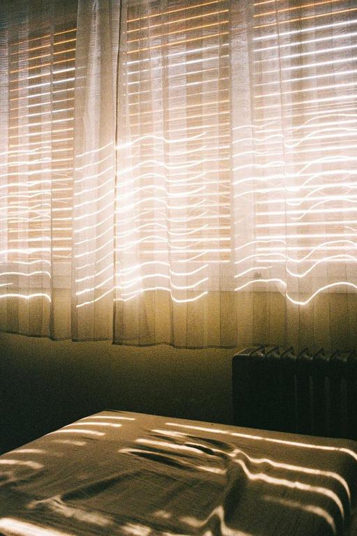 Sunlight streams through window blinds and curtains, casting patterns on a bed and wall.