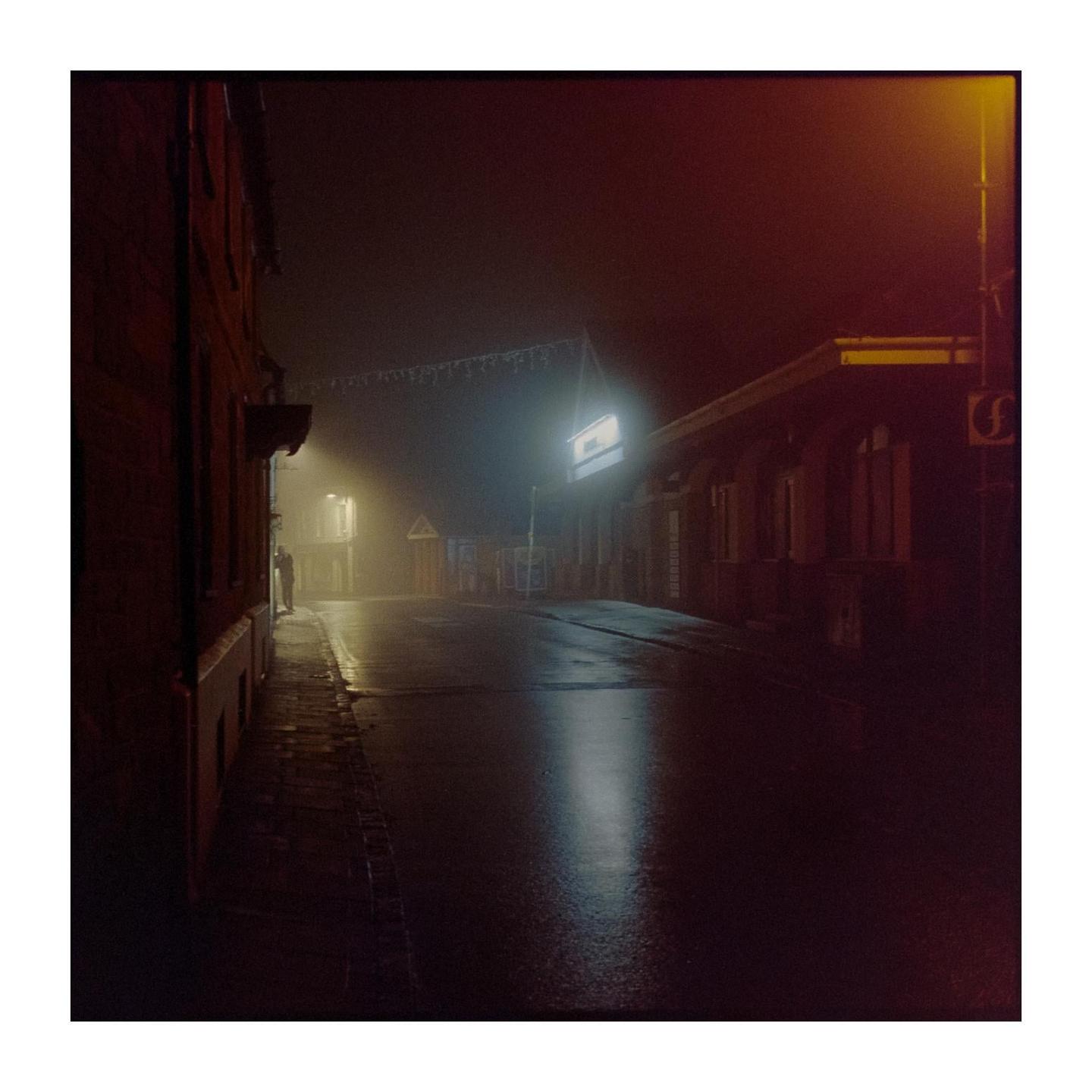 A street at night is illuminated by a bright light and a streetlamp, with a person standing in the distance.