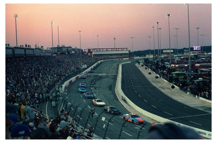 Race cars speed around a track at dusk, surrounded by a large crowd in the stands.