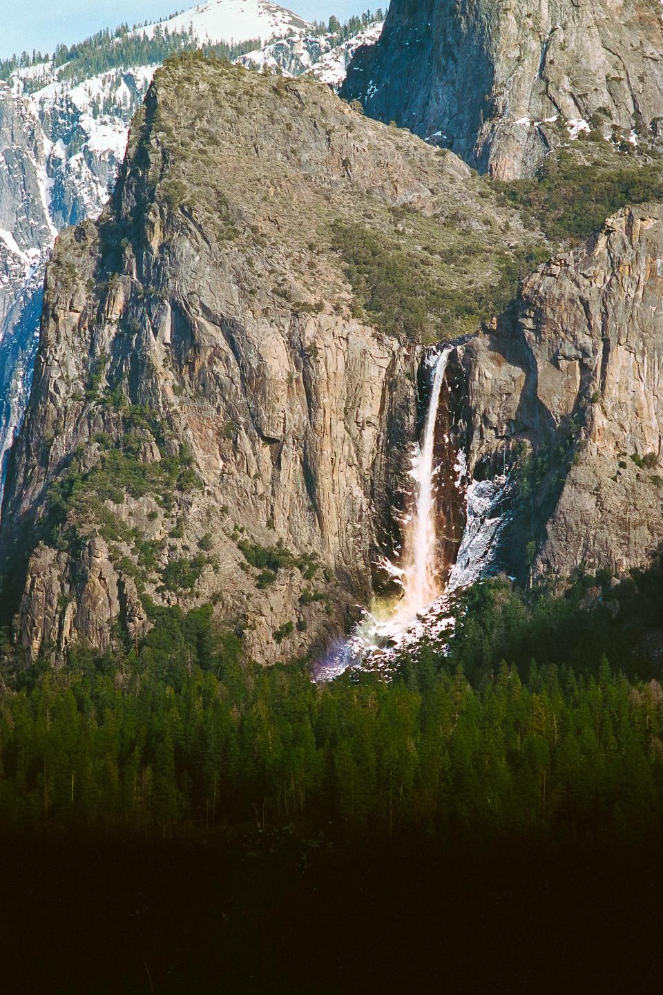 A waterfall cascades down a rocky cliff face, with a rainbow visible at its base and a forest of green trees below.