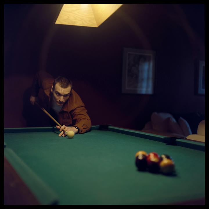 A man in a brown jacket leans over a pool table, aiming a cue stick at the cue ball.