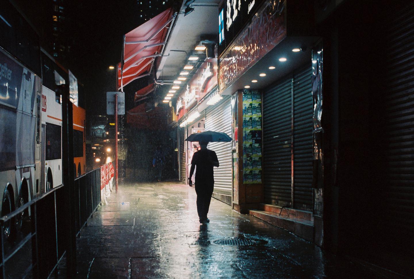 A person walks with an umbrella down a wet street at night, illuminated by storefront lights and passing vehicle headlights.