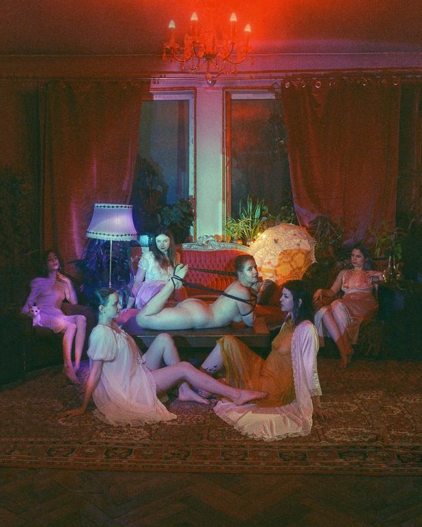 Six women and a cat are in a dimly lit room with red and blue lighting.