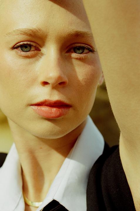A woman with light skin and light hair looks directly at the viewer, with a shadow cast across her forehead and a bright highlight on her cheek.