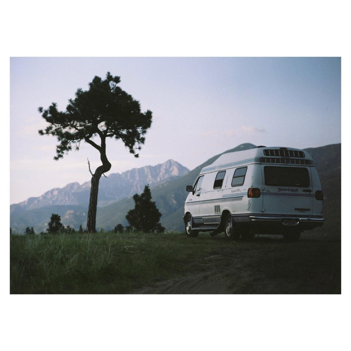 A white van is parked on a grassy hill with mountains in the background and a tree to the left.
