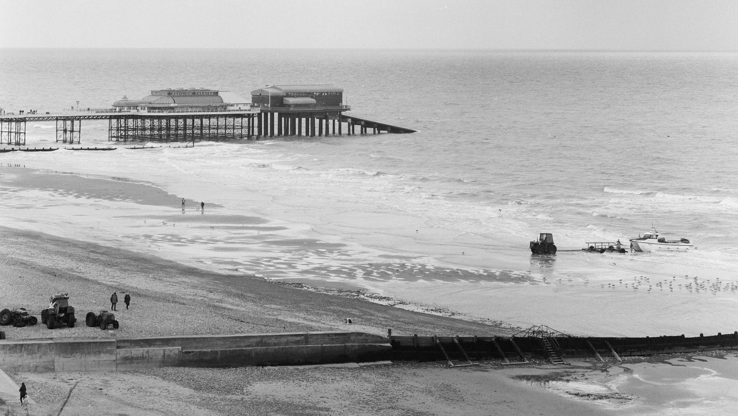 A pier extends into the ocean next to a beach with a tractor pulling a boat into the water.