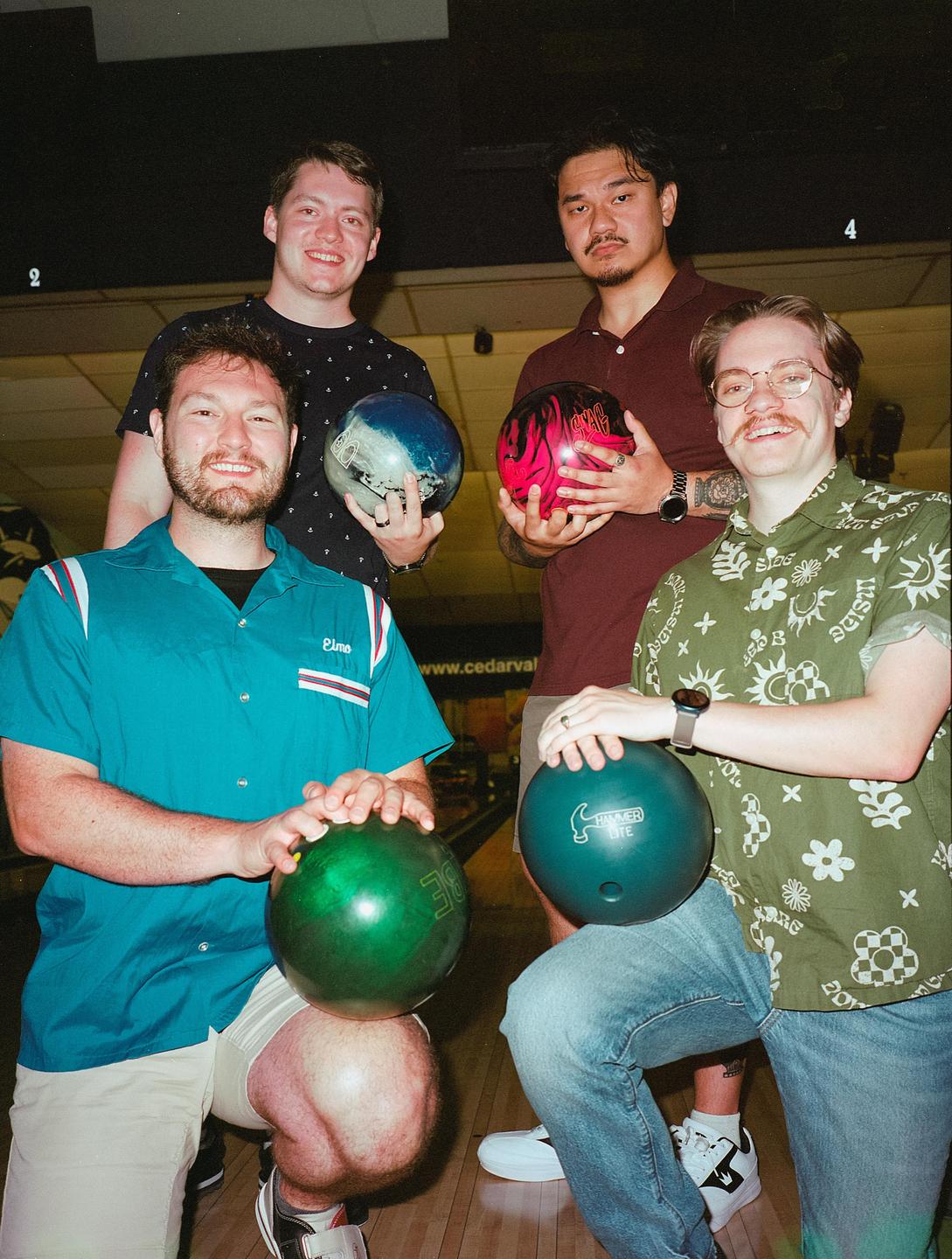Four men pose with bowling balls at a bowling alley.