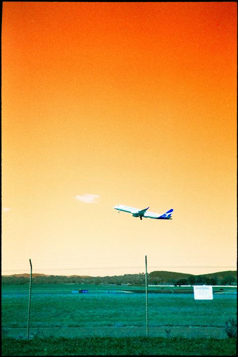 An airplane takes off over a green field with a fence in the foreground under an orange sky.