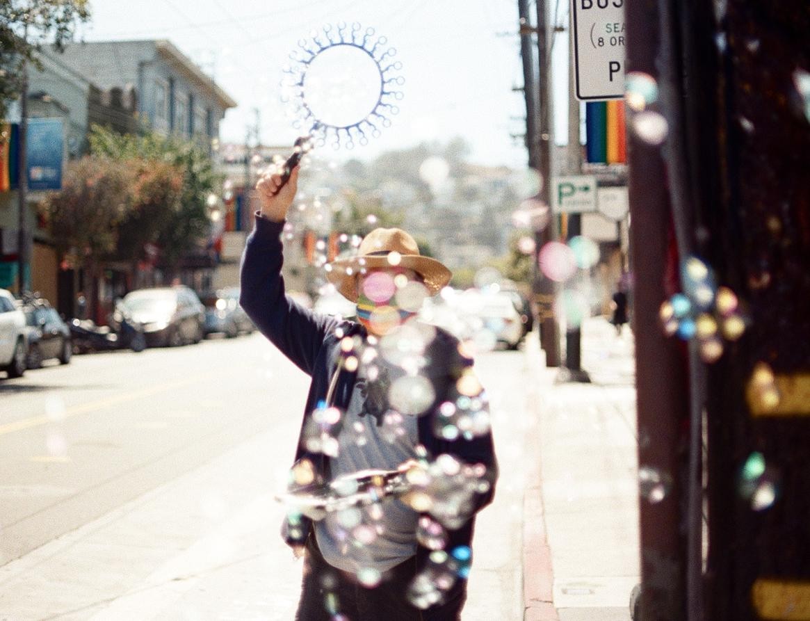 A person in a straw hat blows bubbles on a sunny street lined with cars and buildings.
