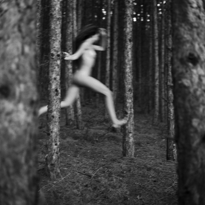 A blurred nude woman runs through a forest of tall trees.